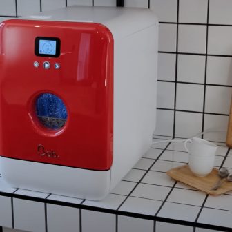 Hyperrealistische liggende foto van een futuristische consumentenelektronicawinkel: een klant loopt door kassaloze poortjes terwijl zij haar smartphone gebruikt, links staat een medewerker met tablet klaar voor ondersteuning en rechts test een andere klant een smartphone; rondom tonen elektronische schaplabels, laptops en grote schermen de high-tech winkelervaring zonder zichtbare teksten in beeld.